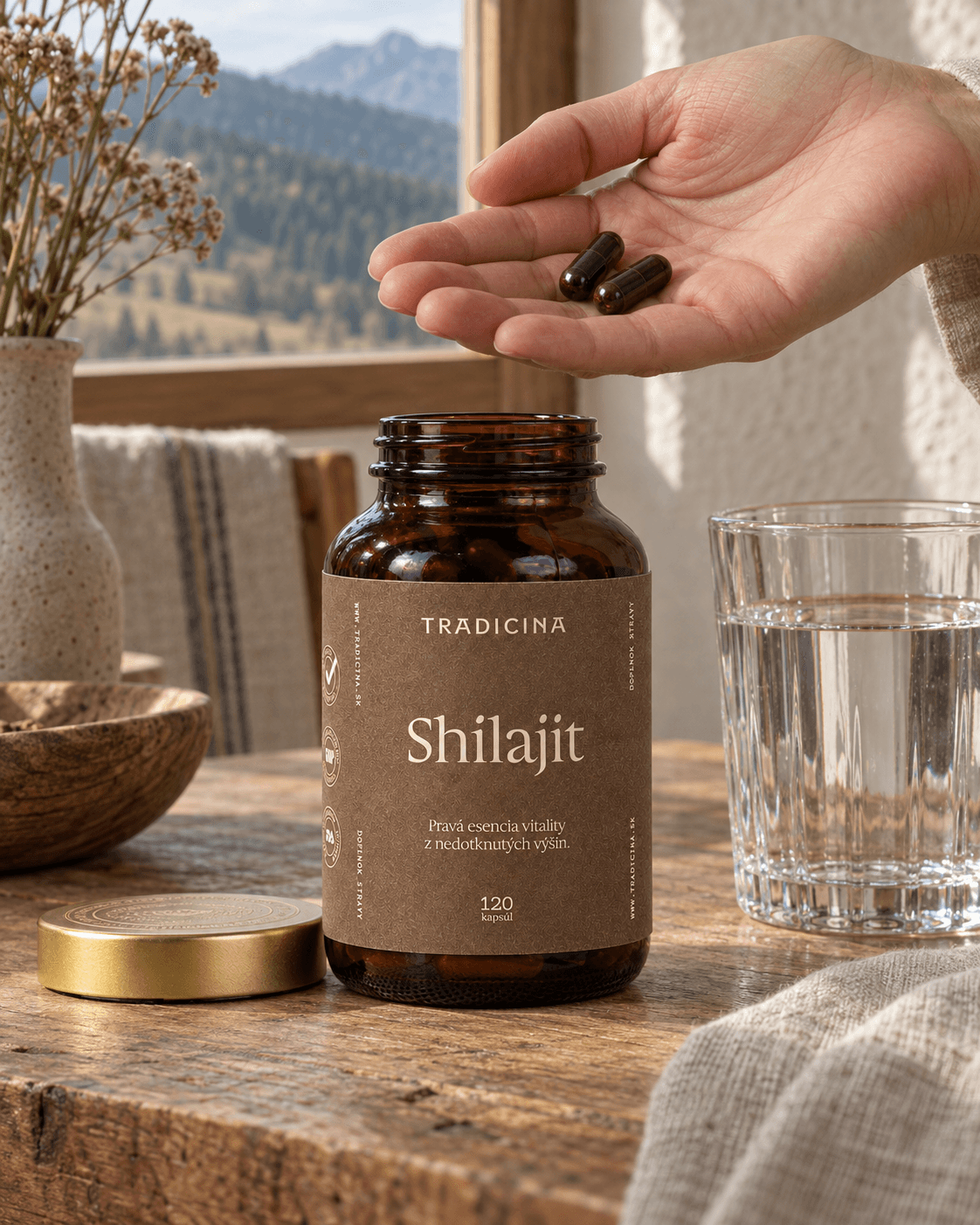 Dlaň s dvoma kapsulami Shilajit nad otvorenou fľaškou Tradicina na drevenom stole, vedľa pohár vody a zlaté viečko