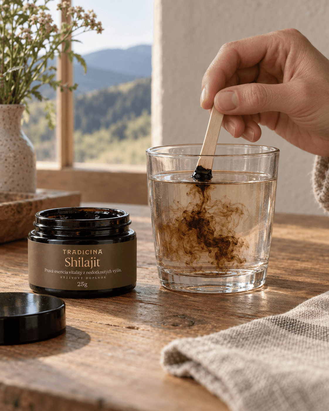 Ruka rozmiešava drevenou lyžičkou kúsok shilajitovej živice v pohári teplej vody, vedľa stojí otvorená dóza Shilajit TRADICINA 25 g